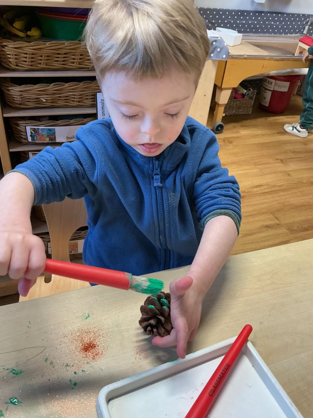 A young boy carefully painting a pine cone at a classroom craft table