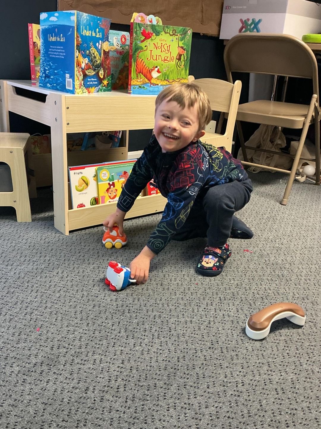A boy laughing while playing with toy cars on the floor next to a children's bookshelf