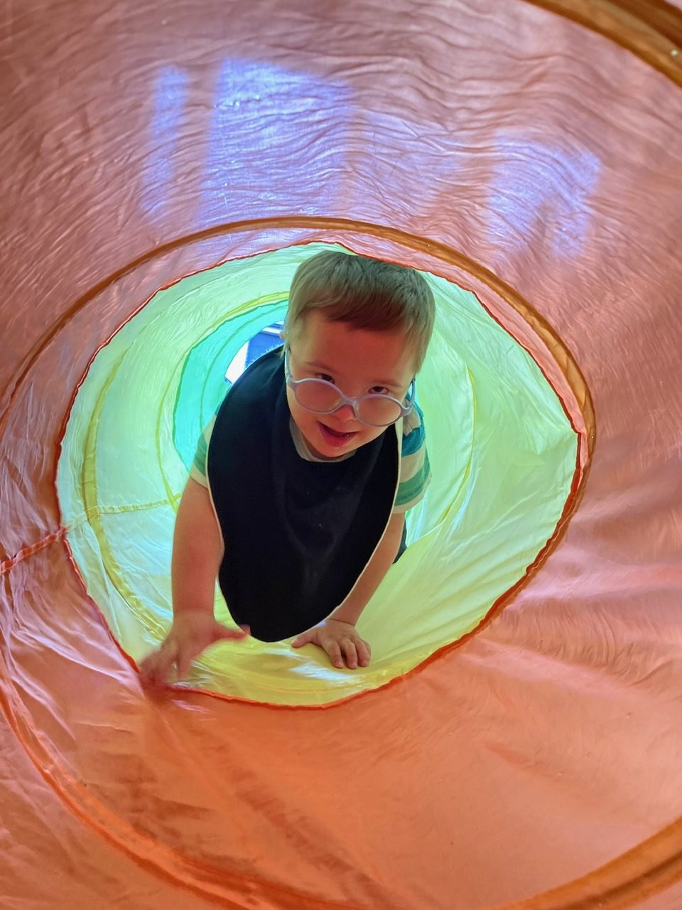 A boy with glasses grinning while looking up from inside a colourful play tunnel