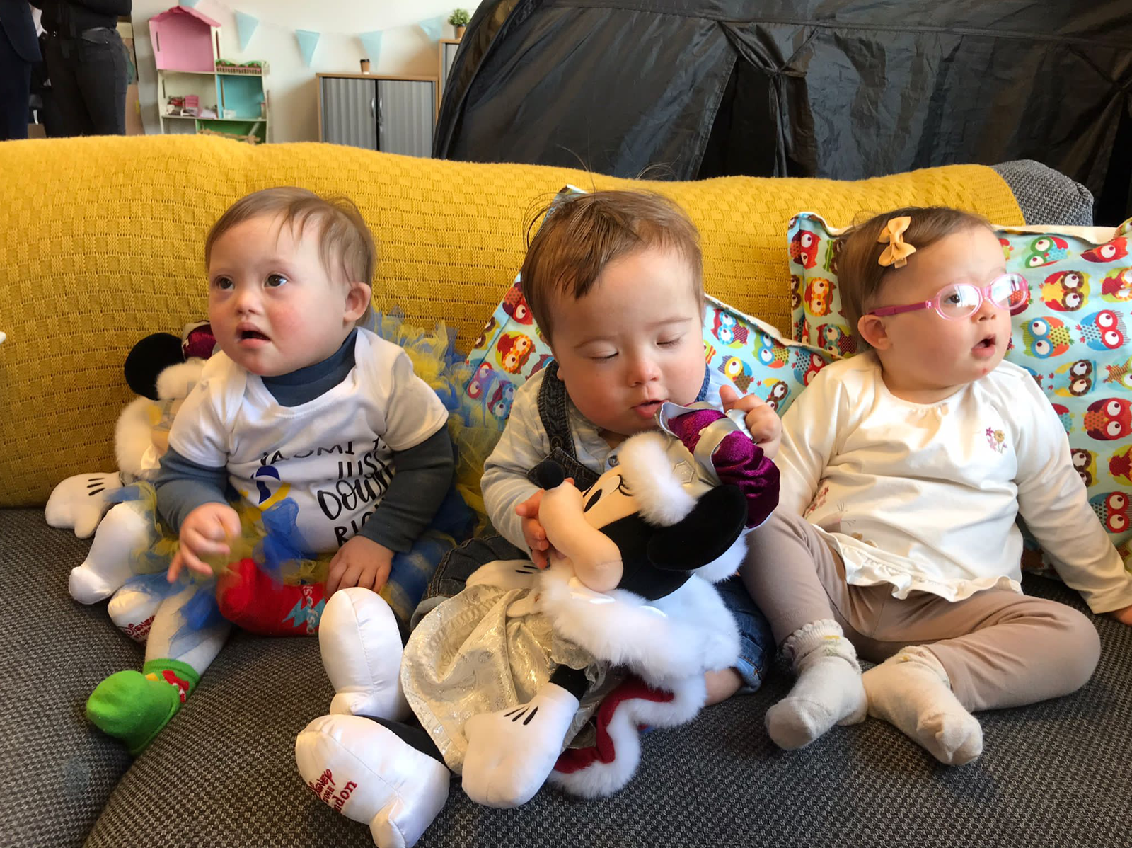 Three babies sitting together on a sofa with cuddly toys