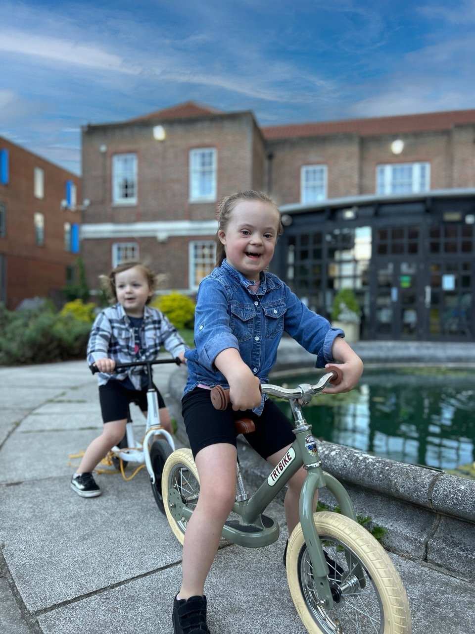 A girl grinning broadly on a balance bike, with a younger child on a scooter behind her