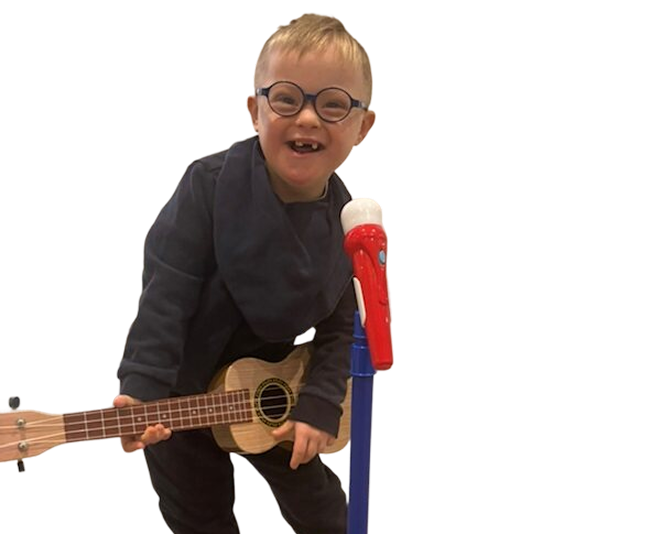 A boy in glasses leaning playfully on a ukulele in front of a toy microphone stand